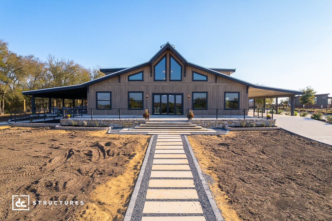 A modern barn-style building with large windows, a wide front entrance, and a stone path leading to the door in a landscaped area.