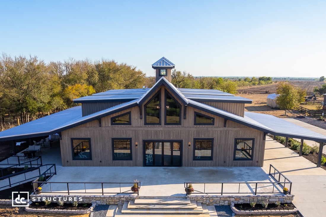A modern barn-style building with large windows, a cupola on the roof, and a spacious patio; surrounded by trees and fields.