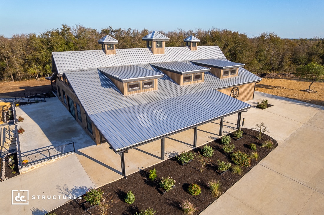 A large modern barn with a gray metal roof, dormer windows, and covered porch sits by gardens and a paved driveway among trees.