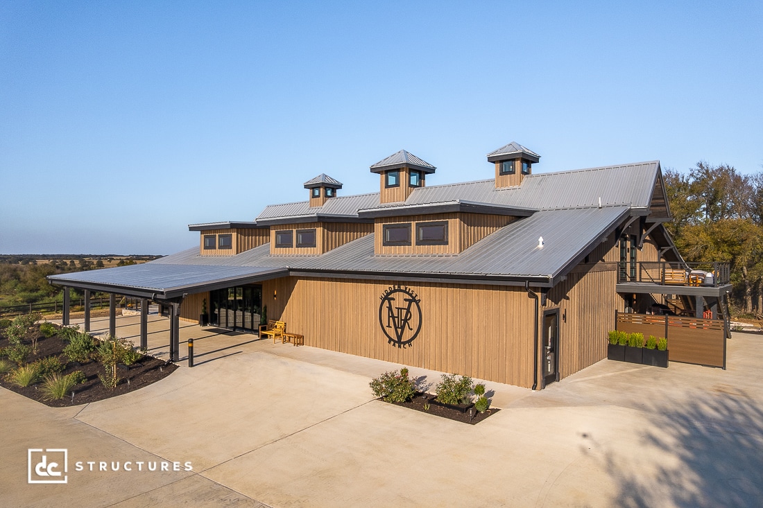A large modern barn-style building with wood siding, metal roofing, and three cupolas, surrounded by greenery and a concrete driveway under a clear blue sky.