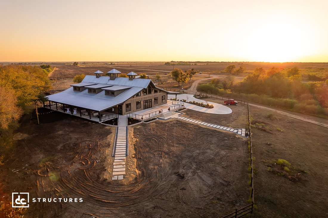 Aerial view of a large modern barn-style building with a gray metal roof, surrounded by dirt and sparse vegetation at sunset. A driveway with a parked red truck leads to the entrance.
