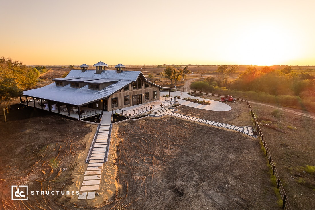 A large, modern barn-style building with a metal roof sits on a spacious rural property at sunset. Concrete walkways and landscaping surround the structure. The sky is clear and golden.