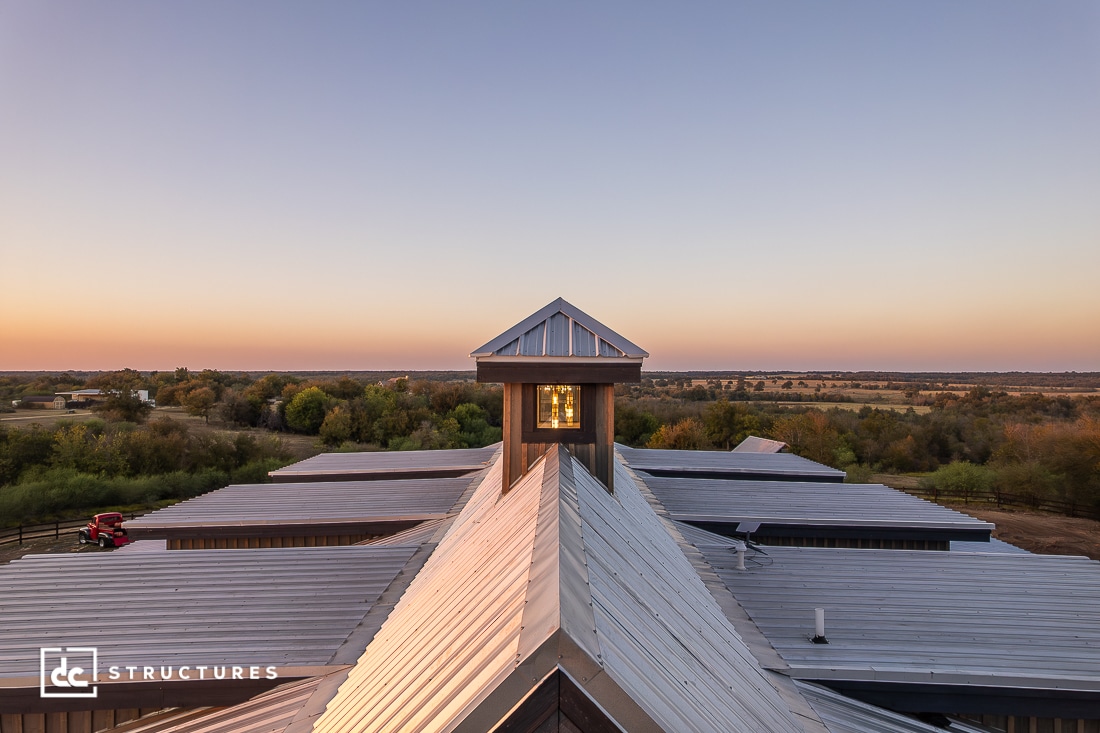 A modern barn with a metal roof and central cupola sits in a rural landscape at sunset, surrounded by fields and trees under a clear sky.