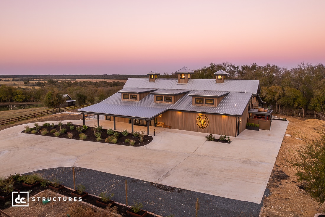 A large modern barn-style building with a metal roof, cupolas, and wraparound porch sits among greenery under a pastel sky.