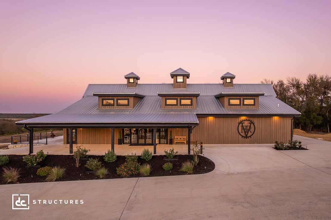 A large modern barn with a metal roof and wooden exterior at sunset, three cupolas, large windows, plants, and concrete driveway.