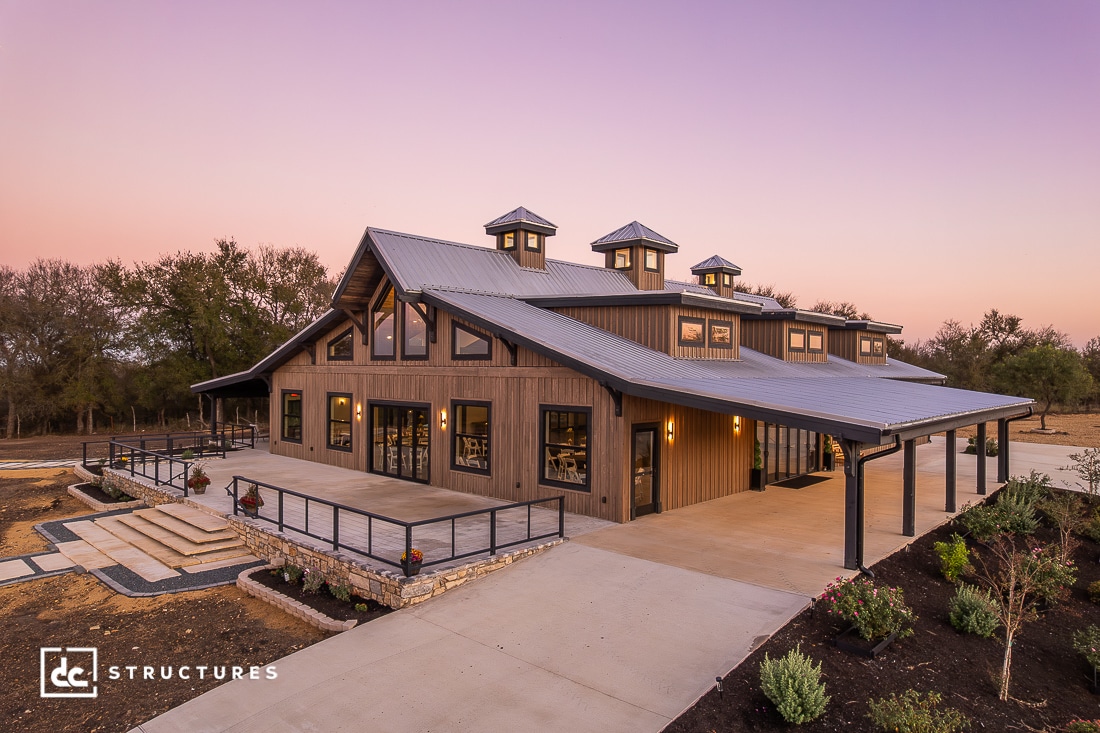 A modern barn-style building with large windows, metal roof, and cupolas, surrounded by landscaped paths and greenery at sunset.