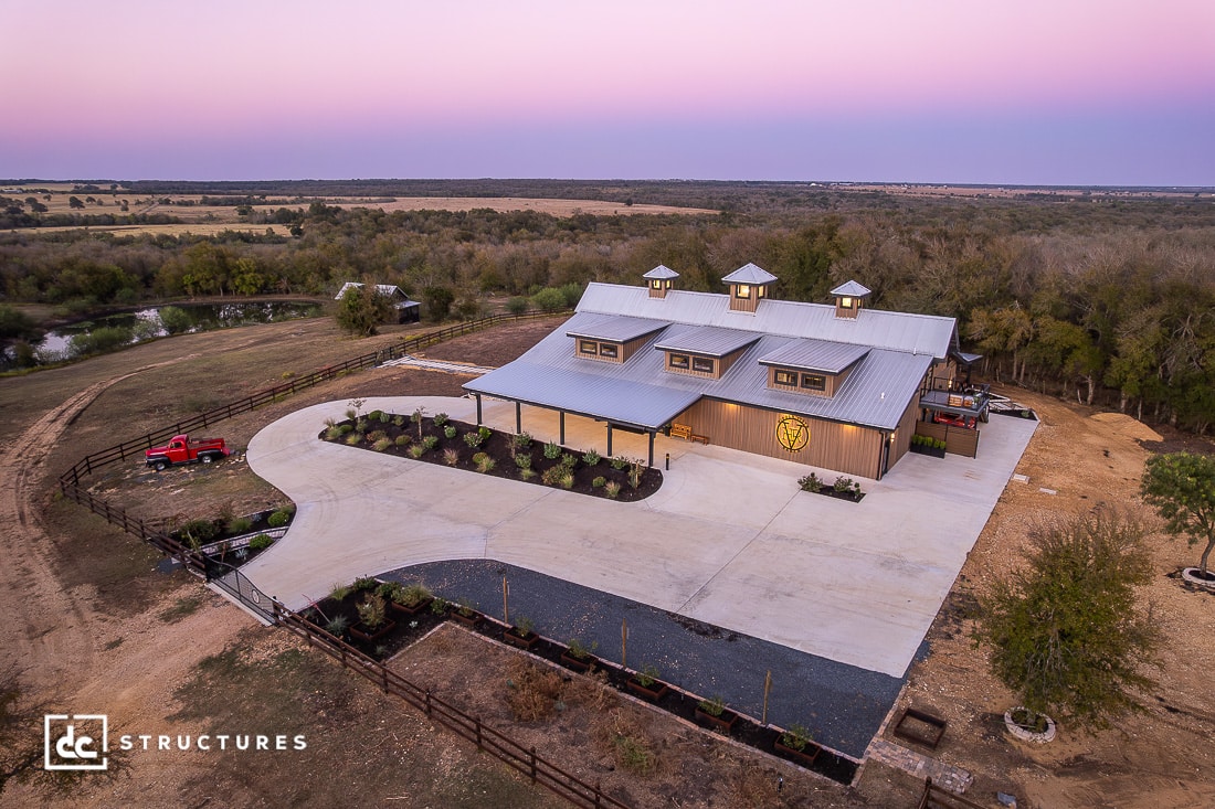 A large modern barn with three cupolas sits on a landscaped property at sunset, with a curved driveway, red truck, and pond nearby.