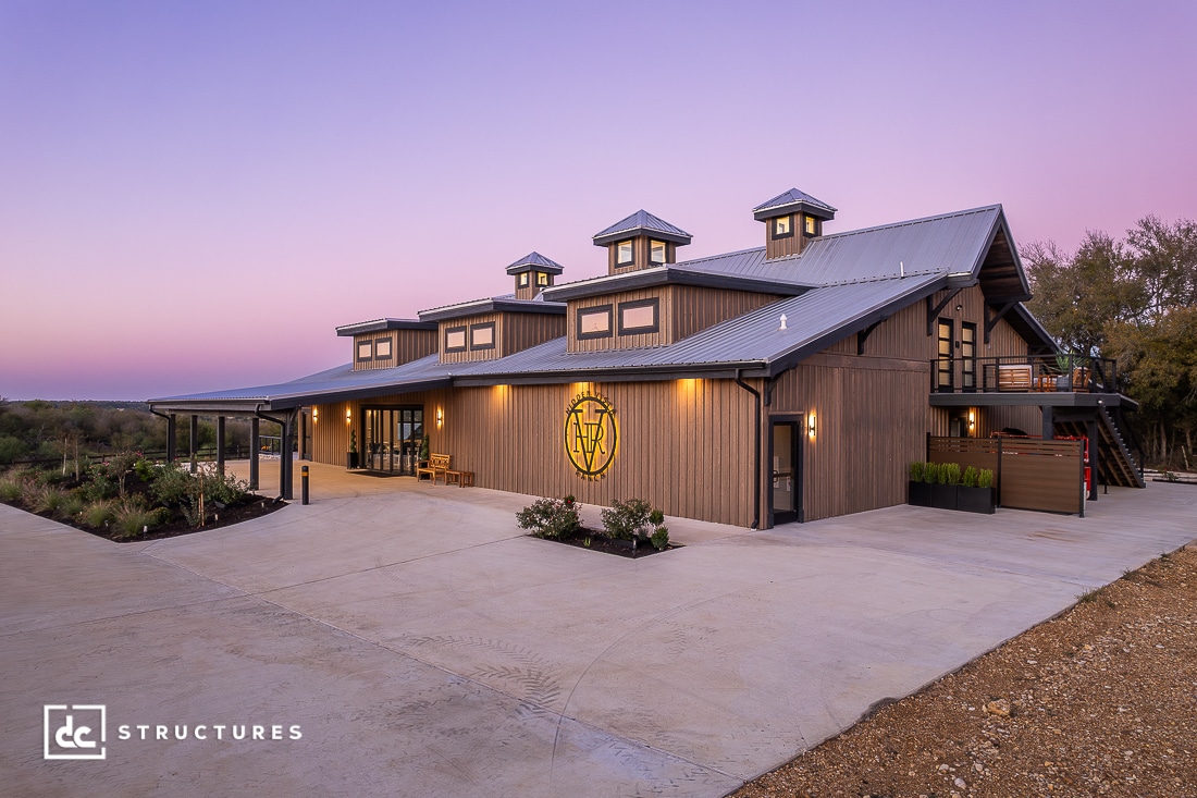 A modern barn-style building with three cupolas, large windows, and wood siding sits on a spacious concrete lot at sunset. The sky is purple.