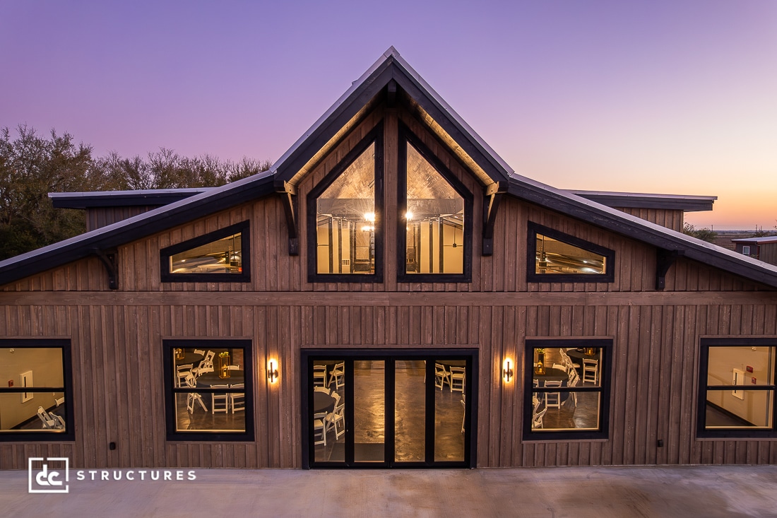 A modern wooden barn with large windows and double glass doors is brightly lit from inside at sunset. Several tables and chairs are visible through the windows. The sky is purple and orange.