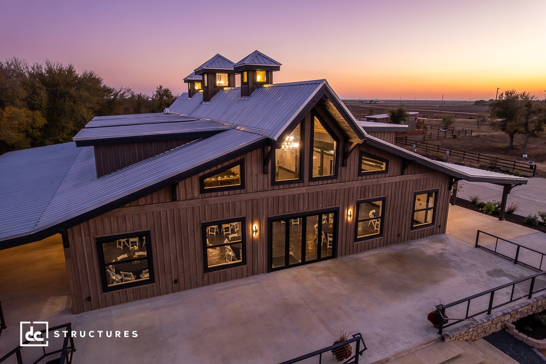 A modern barn-style building with large windows and a metal roof, warmly lit at sunset under a purple-orange sky with trees and fields.