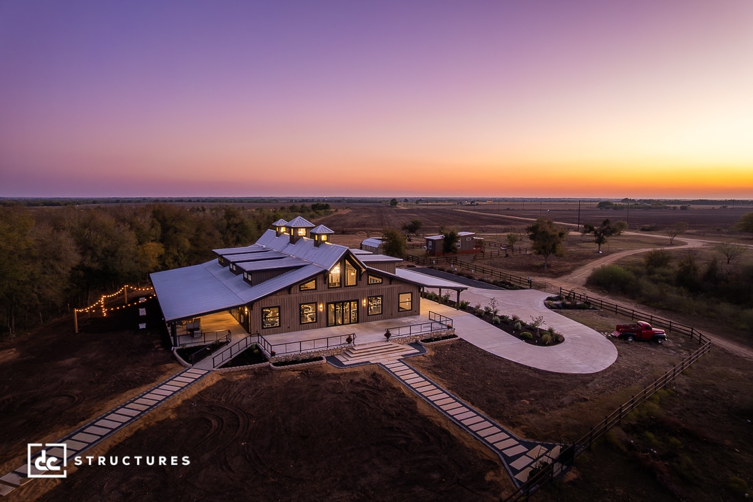 A modern barn-style home with large windows and exterior lights sits on a rural property at sunset, surrounded by trees and a pickup truck.