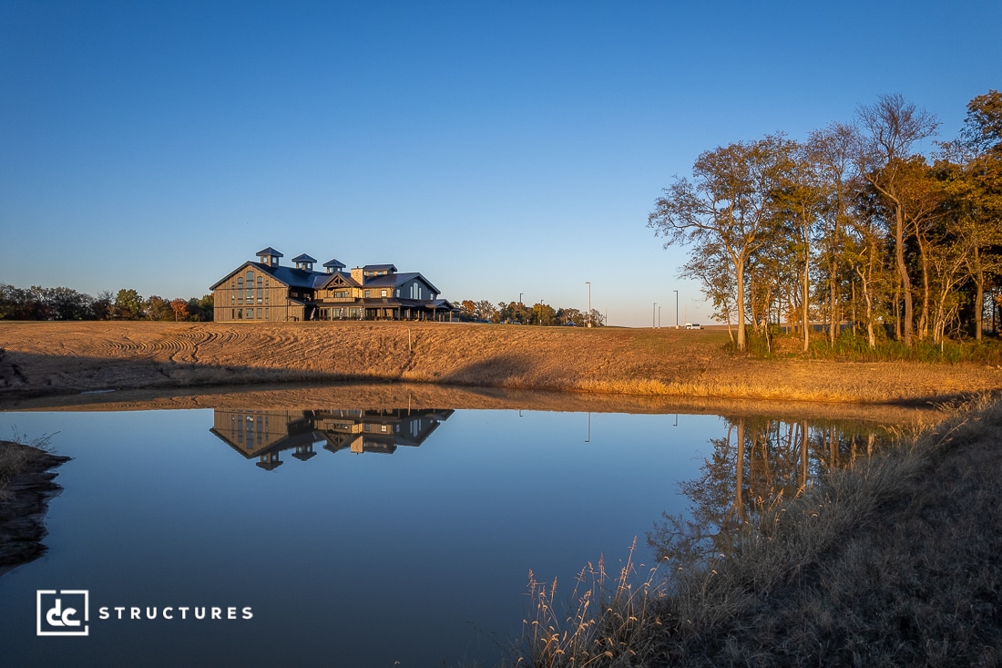 A large lodge-style building sits atop a hill, reflected in a calm pond below. Trees with autumn foliage line the right side, and the sky is clear and blue.