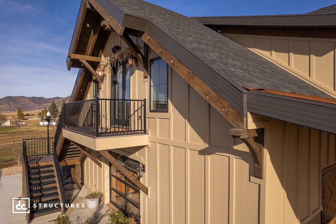 A modern barn-style building with beige vertical siding, dark wood trim, a balcony, exterior staircase, and mountains behind.