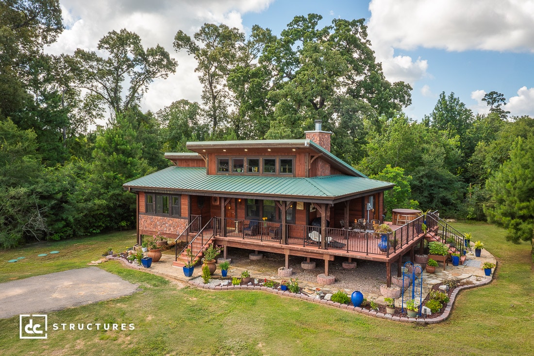 A rustic wooden house with a green metal roof and wraparound porch sits on a grassy lot, surrounded by trees and potted plants. The sky is partly cloudy.