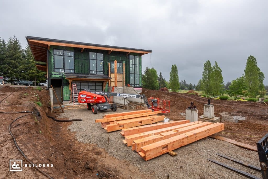 A modern two-story house under construction with large glass windows. Tools and equipment are scattered on gravel in front. Trees and a cloudy sky are in the background.