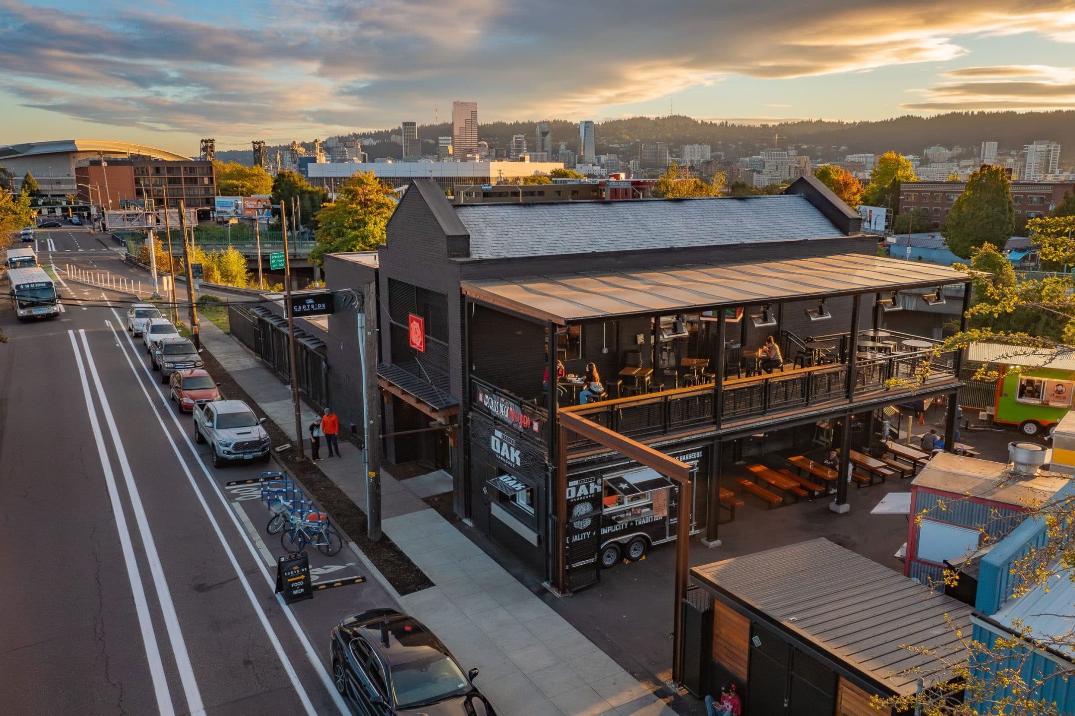 garden-tavern-pdx A two-story modern black building with outdoor seating on the upper level, cars parked along the street, and a city skyline and hills in the background at sunset.