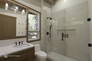Modern bathroom with a glass shower enclosure, wooden vanity, white countertop, wall-mounted mirror, toilet, and a window showing trees. Black fixtures and a sleek, contemporary style.