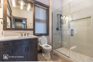 Modern bathroom with dark wood vanity, white countertop, rectangular mirror, toilet, and glass walk-in shower with black fixtures.