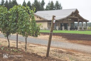 A modern barn-style building stands behind a row of grapevines on a farm, with a gravel path and trees in the background. The sky is overcast.