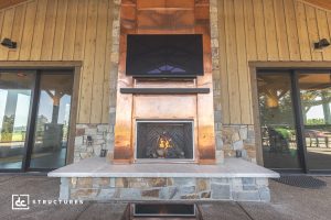 Outdoor stone and wood patio with a copper-framed fireplace burning, large flat-screen TV above, and sliding glass doors on both sides.