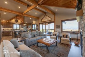 Spacious living room with exposed wood beams, large windows, gray sofas, patterned armchairs, leather ottoman, open to modern kitchen and dining area with scenic views outside.