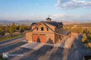 A large, rustic barn-style building with three garage doors sits in a rural landscape surrounded by trees and mountains under a partly cloudy sky.