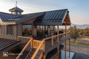 A modern two-story house with wood siding, large windows, a balcony with seating, and a black metal roof in a rural setting.
