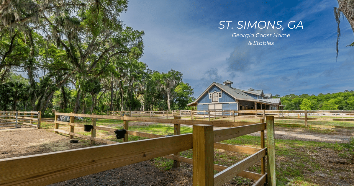 A wooden-fenced horse stable sits beneath tall mossy trees with a blue barn in the background. The sky is partly cloudy.