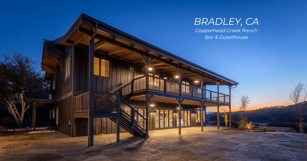Two-story wood guesthouse at sunset with warm lights inside, large wraparound deck, and outdoor stone patio.