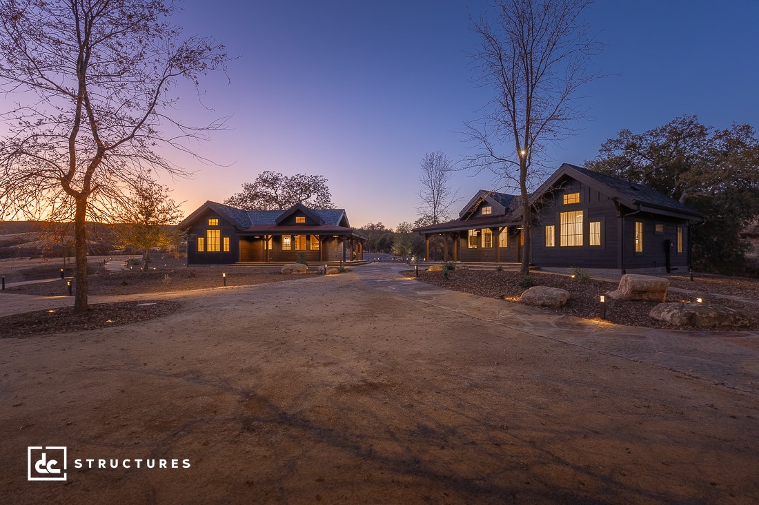 Two modern, single-story houses with large windows are lit at dusk, separated by a dirt path and surrounded by bare trees and rocks.