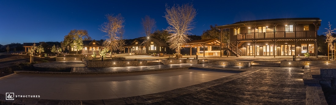 A wide-angle night view of a large, modern house estate with illuminated windows, leafless trees, landscaped walkways, and an empty pool. The deep blue sky indicates dusk or early evening.
