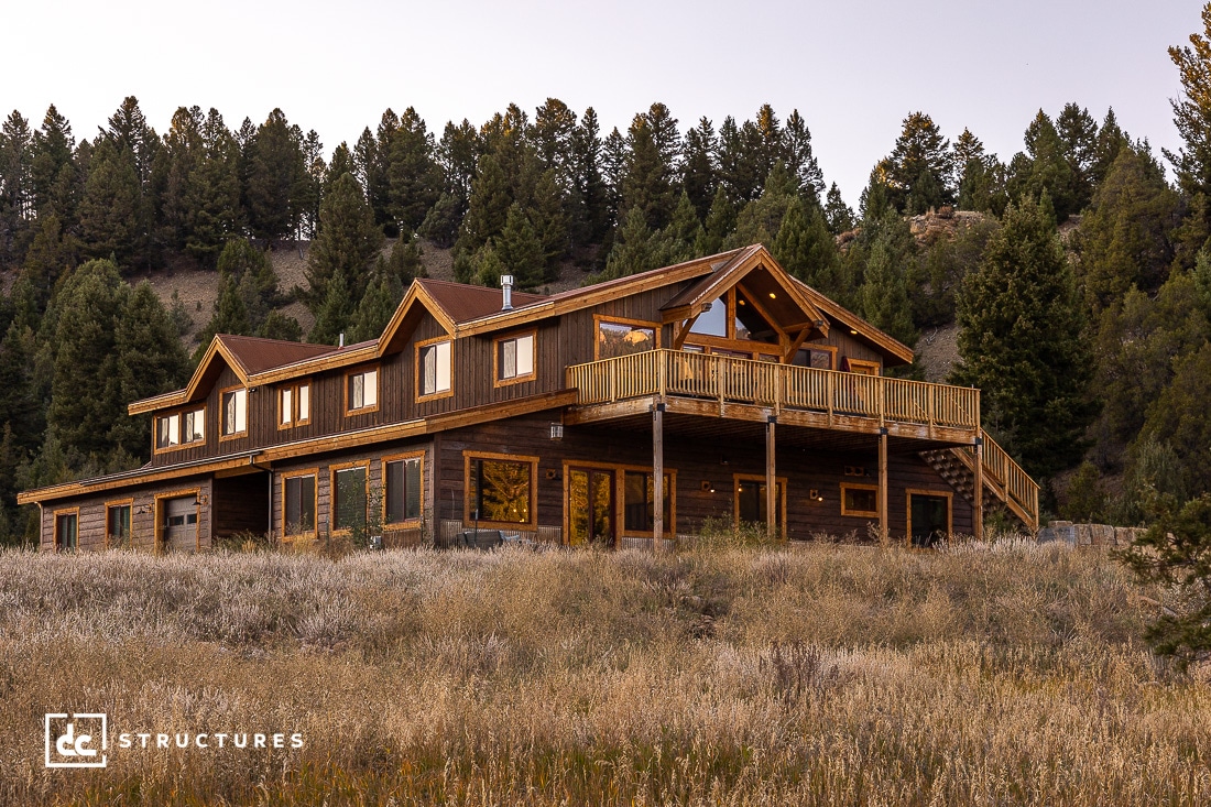 Large two-story wooden cabin with a wraparound deck, tall pine trees and grass, hillside at dusk, multiple windows and gabled roofs.