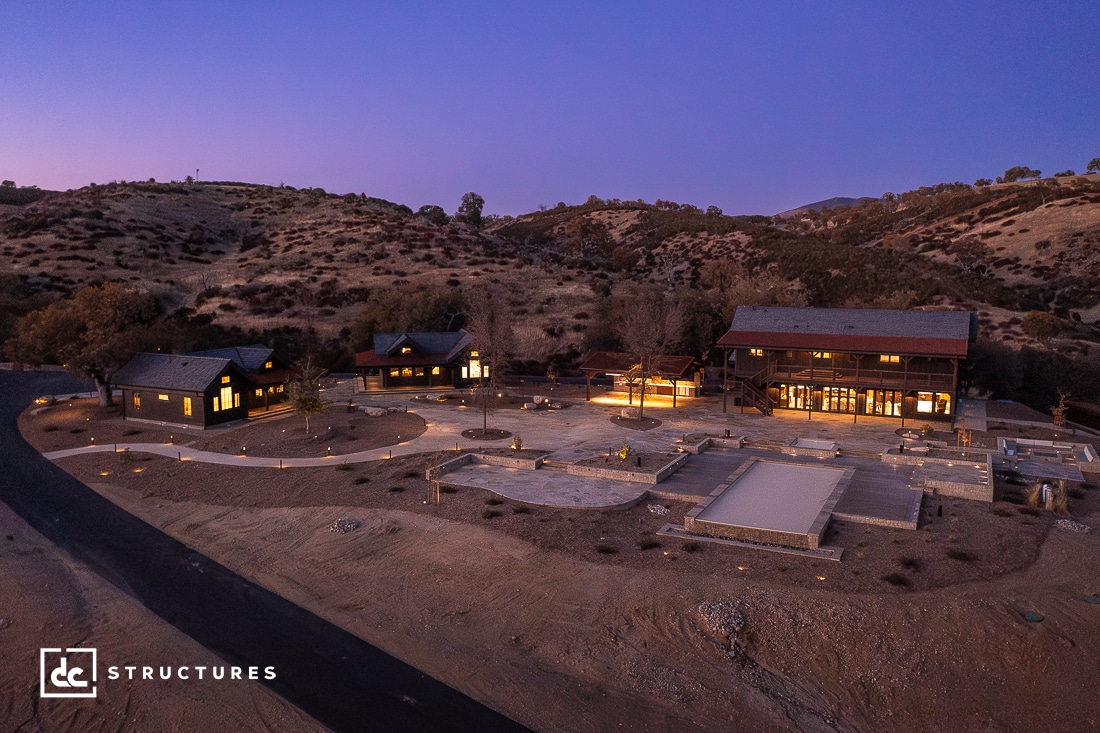 Aerial view at dusk of a modern ranch property with illuminated buildings, paved walkways, landscaped areas, and surrounding hills.