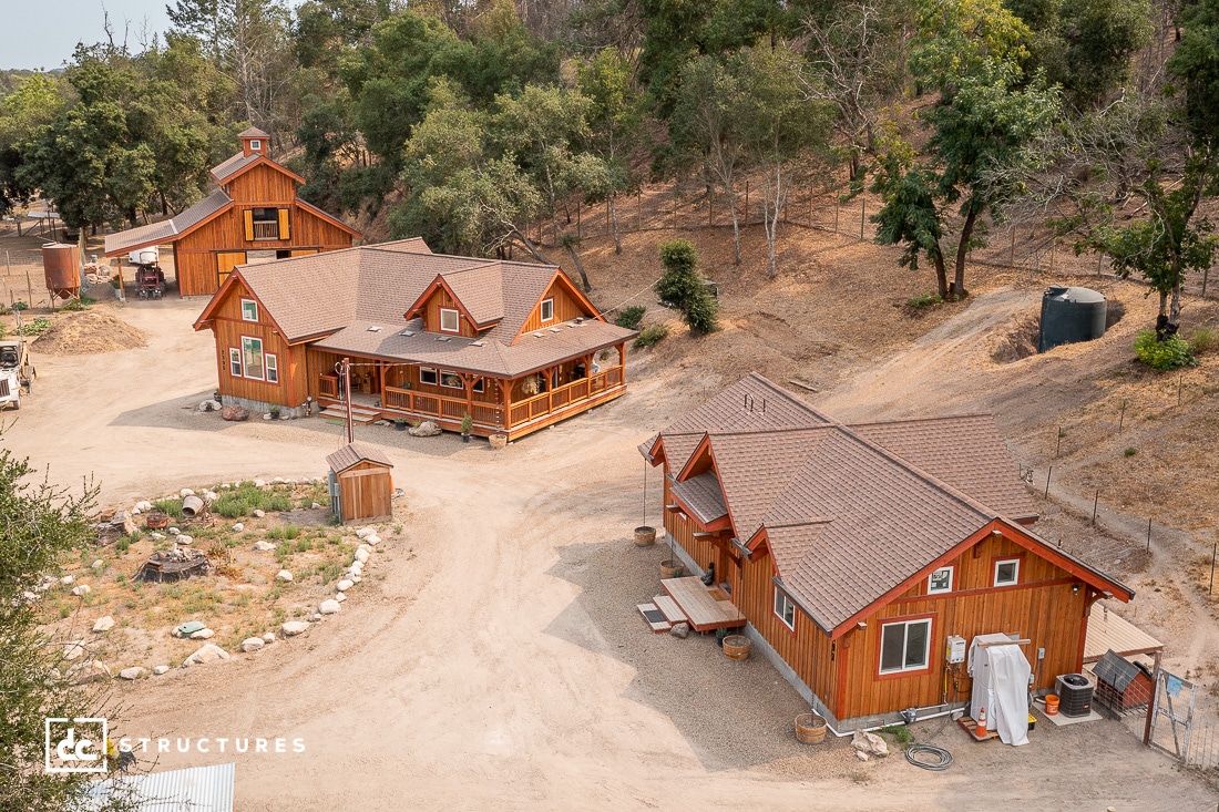 A cluster of three wooden cabins with brown roofs on a dirt driveway, surrounded by dry soil, rocks, trees, a water tank, and shed.
