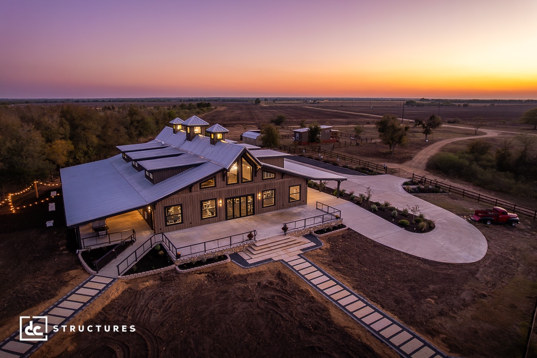 A large modern barn-style building with a metal roof is lit up at dusk, surrounded by paths, landscaping, and open fields. The sky glows with a purple and orange sunset.