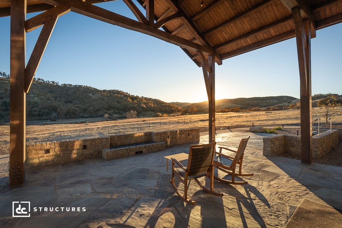 Covered patio with two wooden rocking chairs facing a scenic view of open fields and rolling hills at sunset, framed by stone walls and wooden beams.