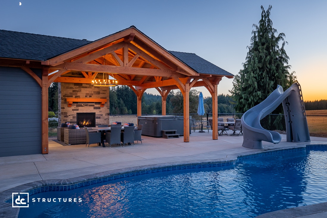 A backyard at sunset with a covered patio, wooden pergola, fireplace, seating, dining area, hot tub, pool with slide, and fields beyond.