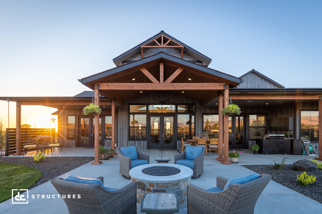 A modern farmhouse with a wooden porch and exposed beams, outdoor seating, a fire pit, and hanging plants at sunset.