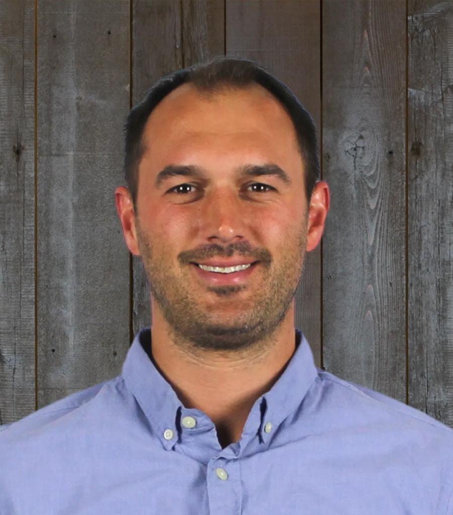 DCS-Vito A man wearing a light blue button-up shirt smiles at the camera. He is standing in front of a wooden plank background.