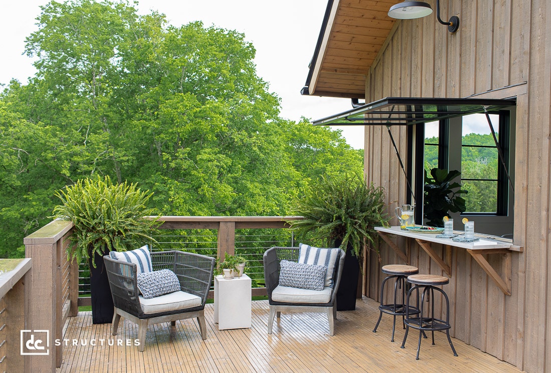A wooden patio with two cushioned chairs, a small table, tall potted plants, and a bar with two stools overlooking green trees.