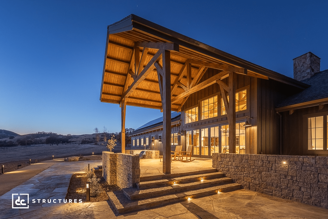 A large modern barn-style building with tall wooden beams, big windows, stone steps, and warm lights at dusk in an open landscape.