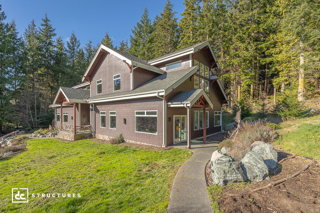 A modern two-story house with dark wood siding and large windows sits among grass, rocks, and tall trees under a blue sky.
