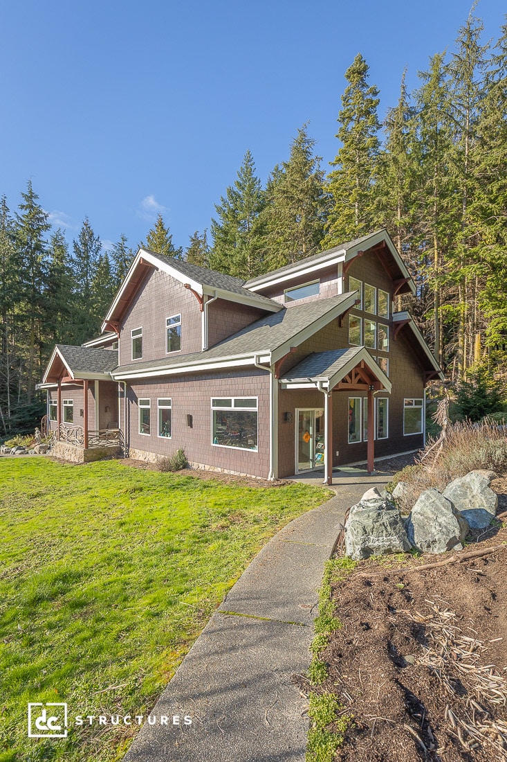 A modern two-story cabin with large windows and dark wood exterior, surrounded by grass and tall pines under a blue sky. Stone path leads to entrance.