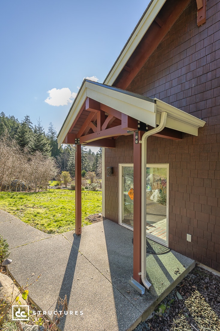 A small covered porch with exposed beams and a metal gutter outside a brown house, leading to a sliding glass door. Sunlit yard nearby.