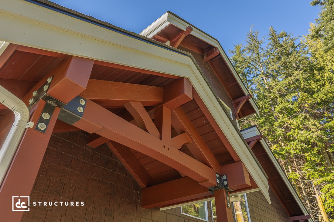 Close-up view of a timber frame gable roof with exposed red wooden beams and metal brackets, set against a blue sky and tall green trees.