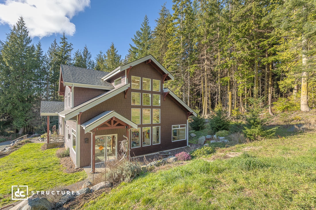 A modern two-story house with large windows sits by a dense forest, featuring a sloped roof, brown siding, and grassy surroundings.