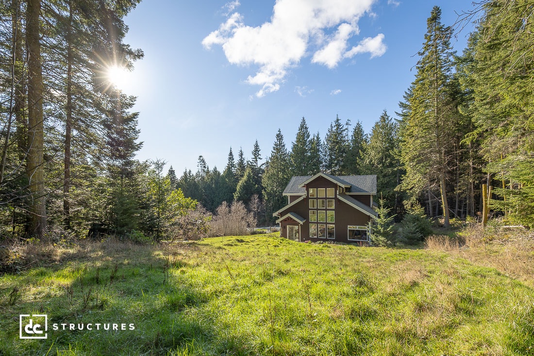 A modern, dark brown wooden house with large windows is nestled in a clearing surrounded by a dense evergreen forest.