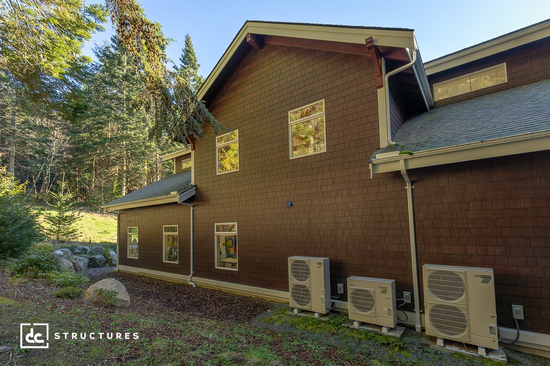 A brown two-story house with several windows sits surrounded by trees. Multiple outdoor HVAC units line the home's exterior wall. The sun is shining.