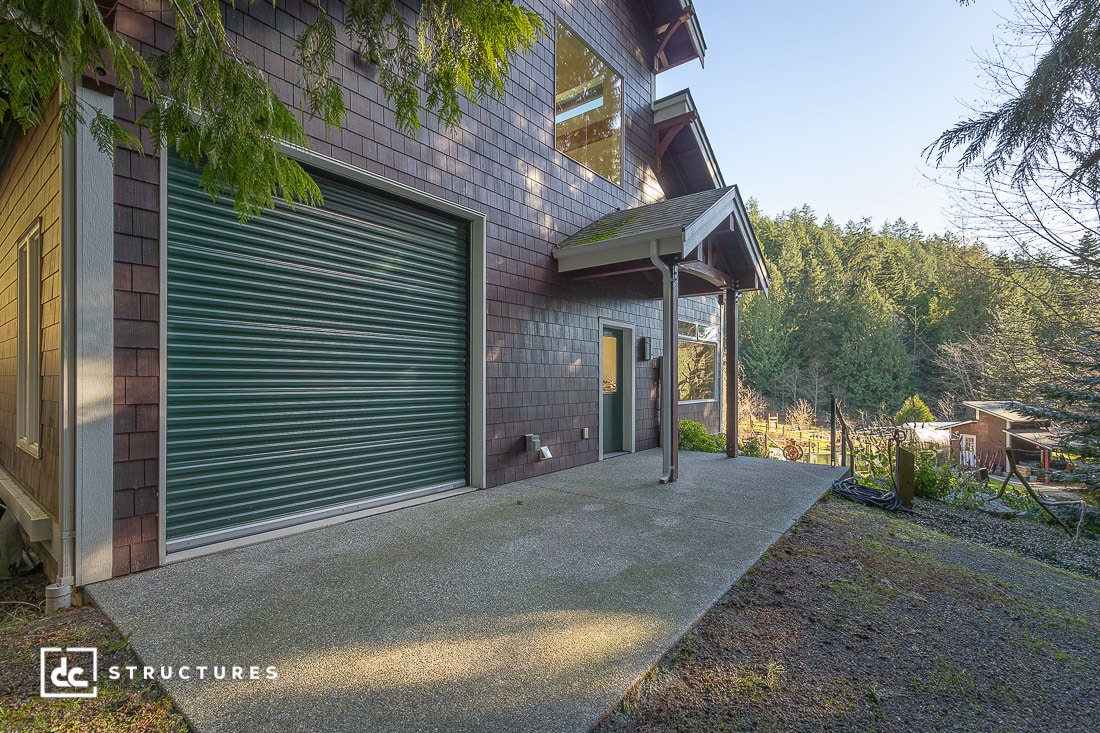 A modern building with brown shingle siding, a large green roll-up door, and glass windows surrounded by trees and a concrete patio on a clear day.