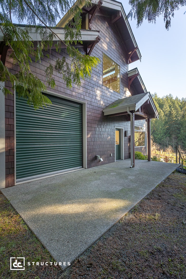 A two-story building with wooden shingles, large windows, a green roll-up garage door, and a small entry door on a concrete patio.